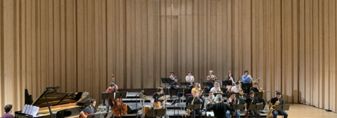 Orquestra de jazz a atuar num palco de auditório, com vários músicos sentados a tocar instrumentos como piano, contrabaixo, bateria e metais, enquanto um maestro dirige o grupo ao centro; ao fundo, uma cortina de madeira clara cobre toda a parede