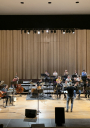 Orquestra de jazz a atuar num palco de auditório, com vários músicos sentados a tocar instrumentos como piano, contrabaixo, bateria e metais, enquanto um maestro dirige o grupo ao centro; ao fundo, uma cortina de madeira clara cobre toda a parede.