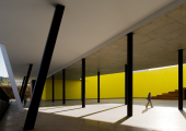 Espaço arquitetónico moderno com colunas inclinadas pretas e uma parede amarela vibrante ao fundo; uma pessoa caminha sob a estrutura com luz natural e sombras marcadas