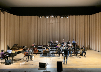 Orquestra de jazz a atuar num palco de auditório, com vários músicos sentados a tocar instrumentos como piano, contrabaixo, bateria e metais, enquanto um maestro dirige o grupo ao centro; ao fundo, uma cortina de madeira clara cobre toda a parede